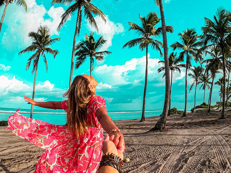 Mulher posando na areia da praia, diante de coqueiros na beira do mar