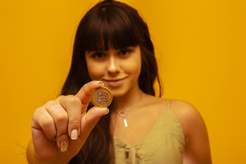 mulher de cabelo longo preto segurando moeda de um real na mão