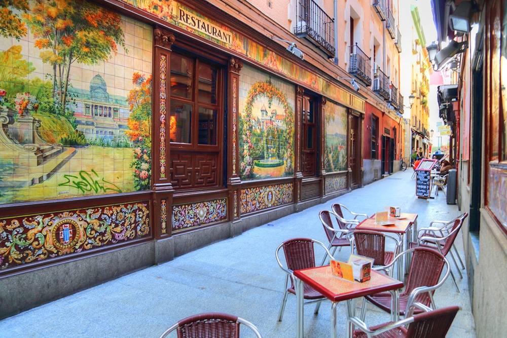 Viela com mesas e cadeiras para refeição de frente com parede de restaurante em azulejos decorados