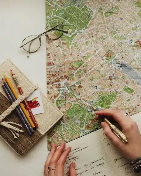 Mapa de papel aberto com óculos de grau, caderno com lápis de cor e mão sobre caderno segurando uma caneta por cima