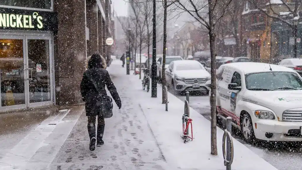 Pessoa caminhando por calçada com carro estacionados na rua ao lado enquanto neva