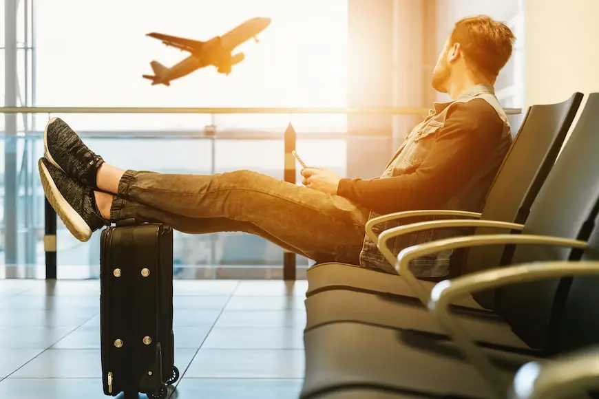 Homem sentado em um saguão de aeroporto, olhando para um avião decolando com as pernas em cima de uma mala