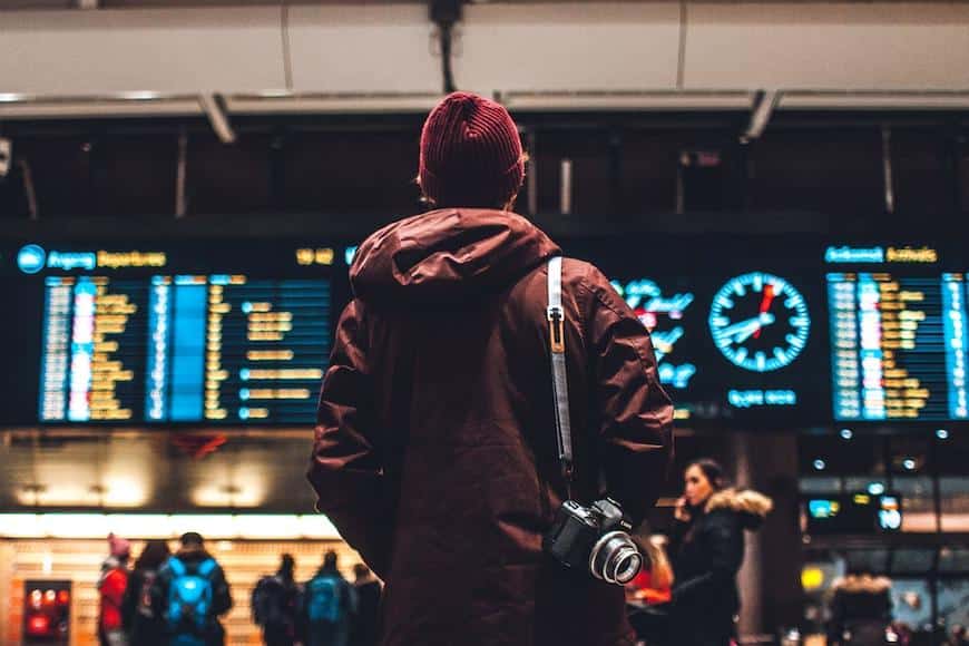 Homem de costas de casaco e gorro cor vinho, uma máquina fotográfica pendurada no ombro olhando para o painel de voos do aeroporto