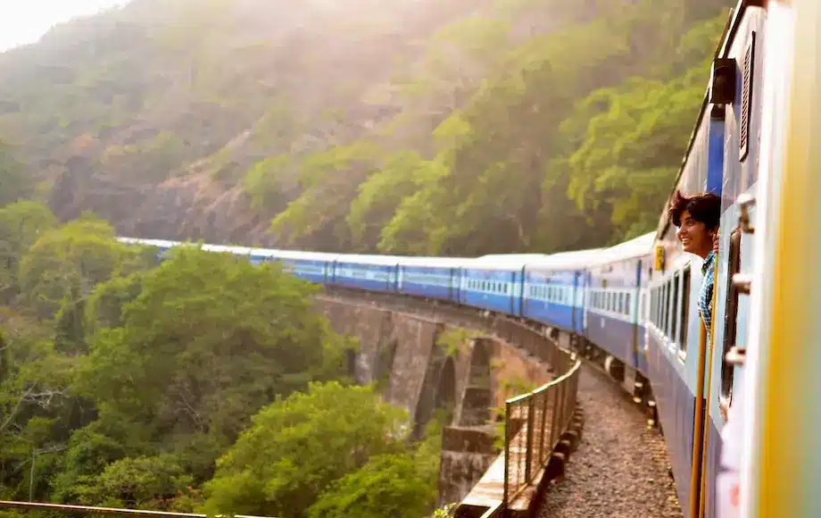 Foto de um trem passando pelo meio de montanhas com uma mulher indiana de cabelo curto sorrindo com a cabeça fora olhando para o horizonte